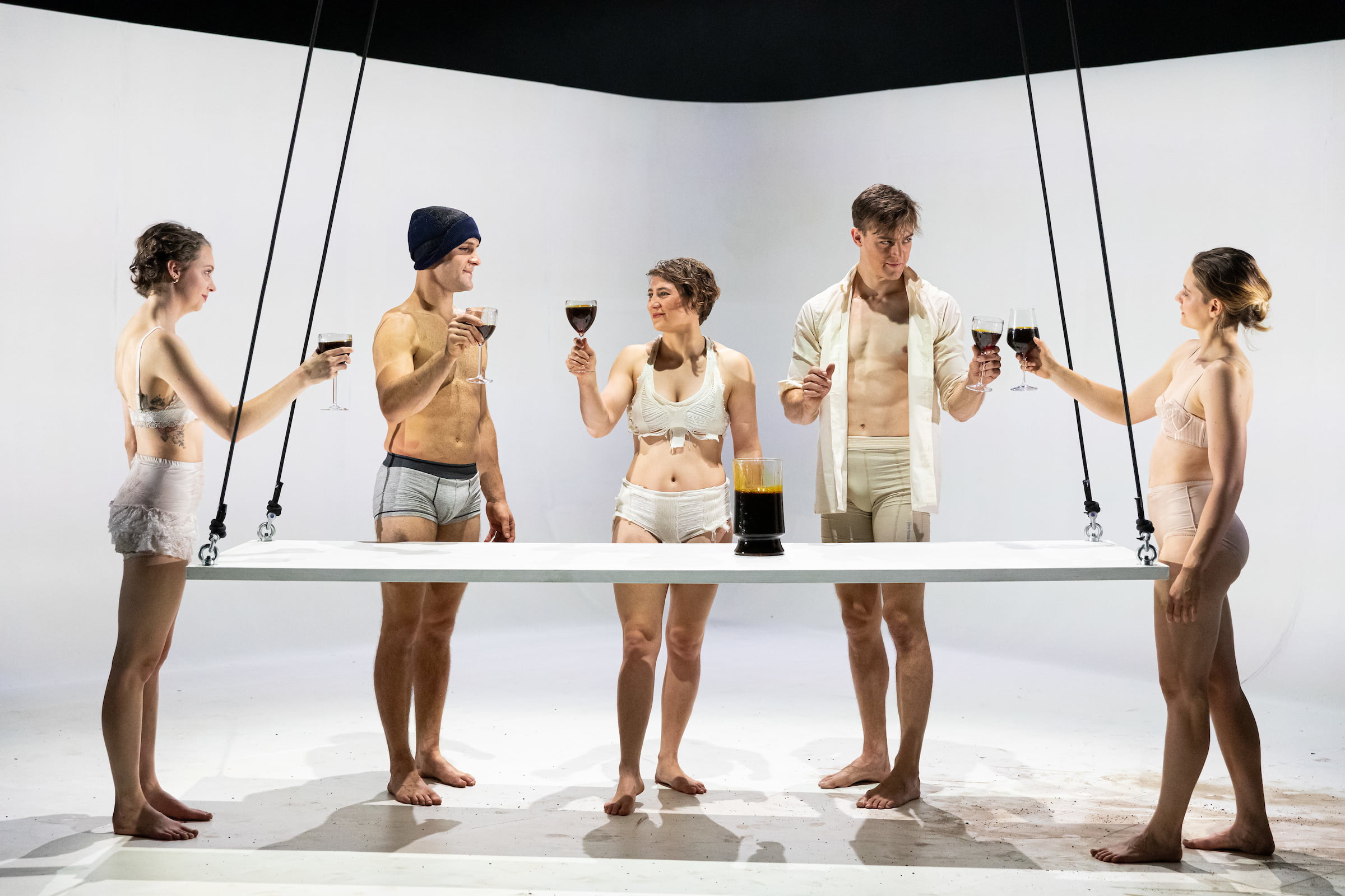 5 performers in nude underwear stand around a blank table and toast each other with wine glasses filled with a dark brown liquid