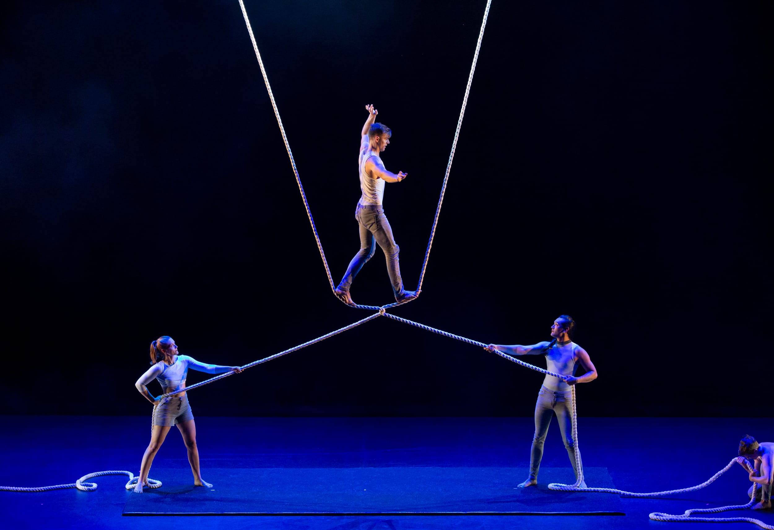 3 performers climb on a structure made of black bars and silver wires in front of a grey building and stone steps