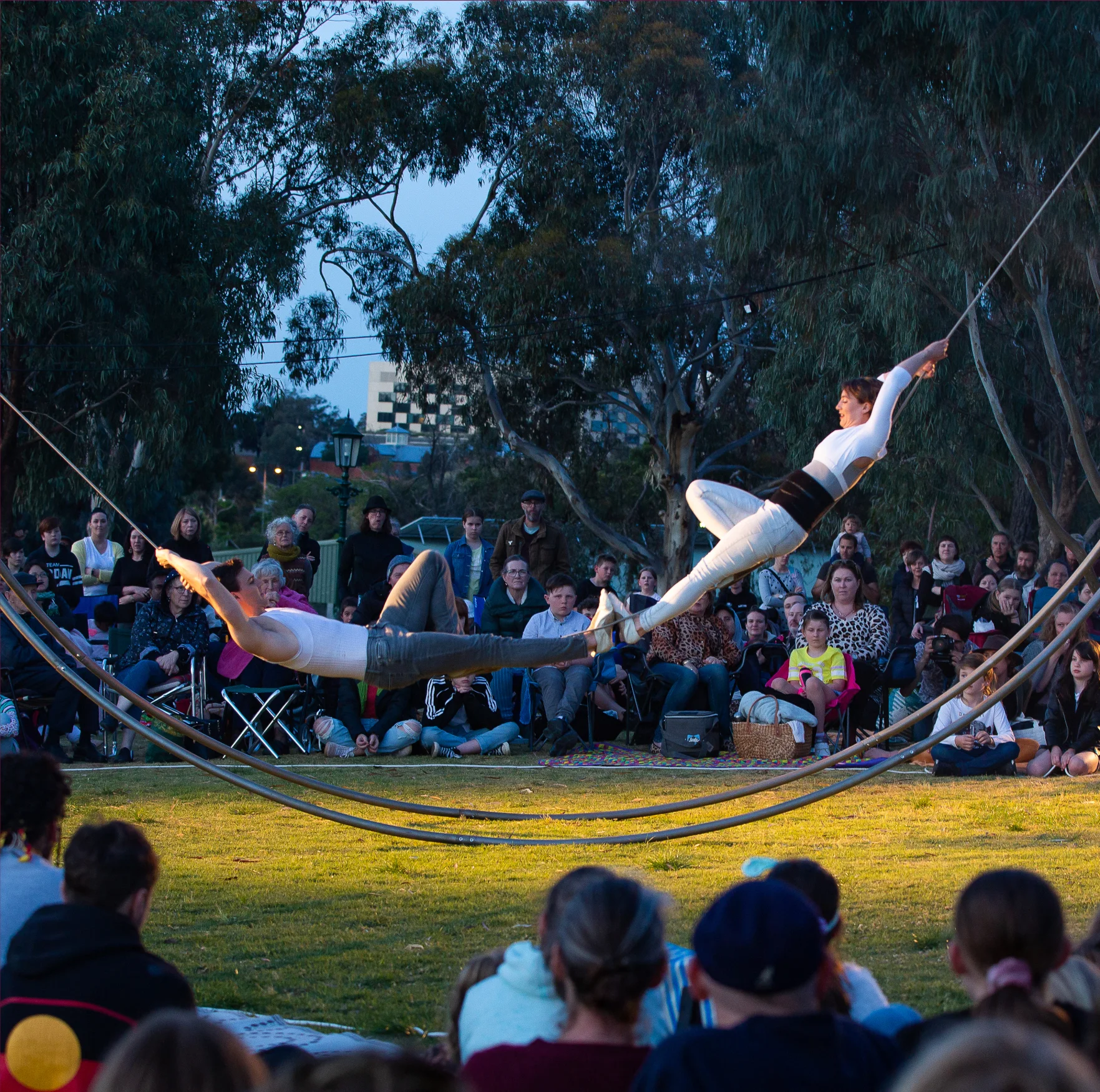 Two performers balance on a rope suspended across a curved metal frame. They are lying down