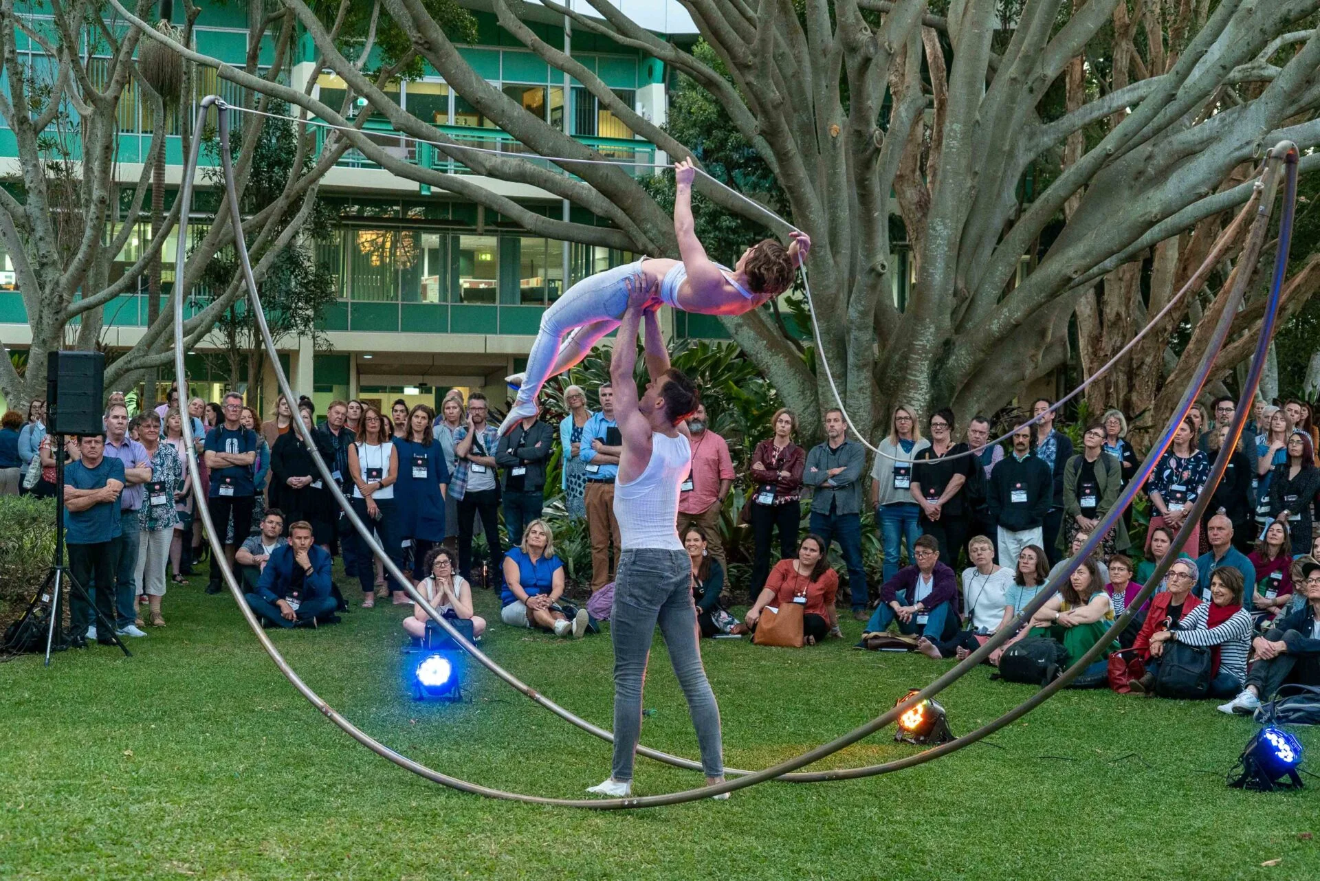 One performer is lifted by another. They are standing inside a curved metal structure. A crowd is watching